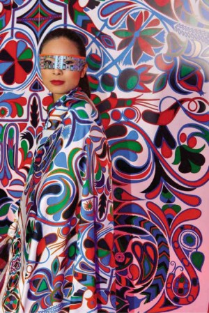 MASKED model draped in an Hermès scarf stands by a photo wall that mirrors scarf’s print.